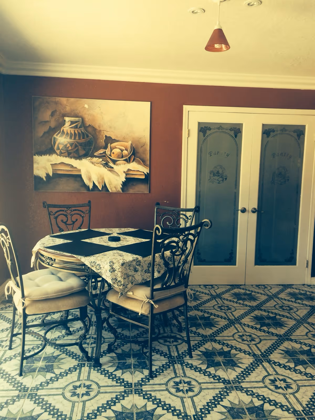 A dining area with a round table covered by a patterned tablecloth and four cushioned chairs with ornate metal backs. The floor has decorative tile with a star pattern. On the wall behind the table is a painting of pottery and fruit. To the right, there are double doors with frosted glass panels labeled 'Pantry'. A single pendant light hangs from the ceiling.