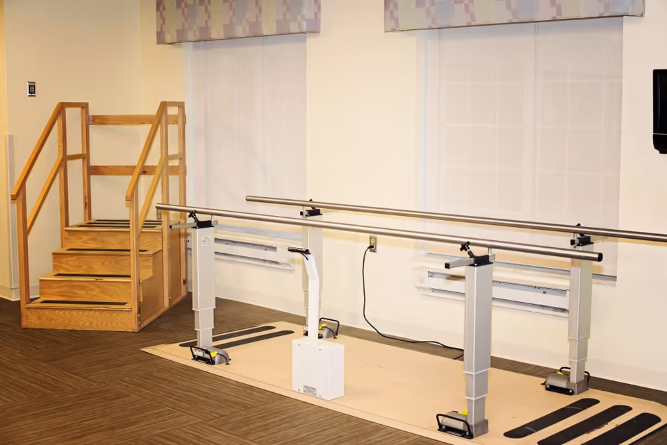 A physical therapy or rehabilitation area with wooden stairs and parallel metal handrails for walking exercises, set against a wall with two windows covered by light-colored blinds.