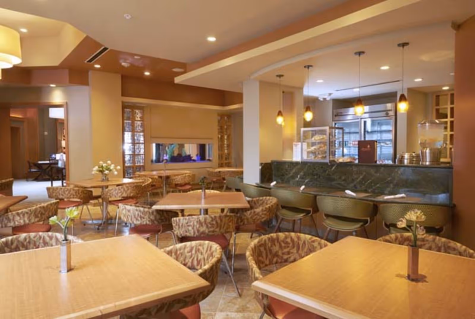 A cozy dining area with multiple wooden tables and patterned cushioned chairs. There are small flower vases on the tables. The room features warm lighting with pendant lights hanging over a counter area that has a display case and a beverage dispenser. The background shows a refrigerator and a fireplace embedded in the wall.