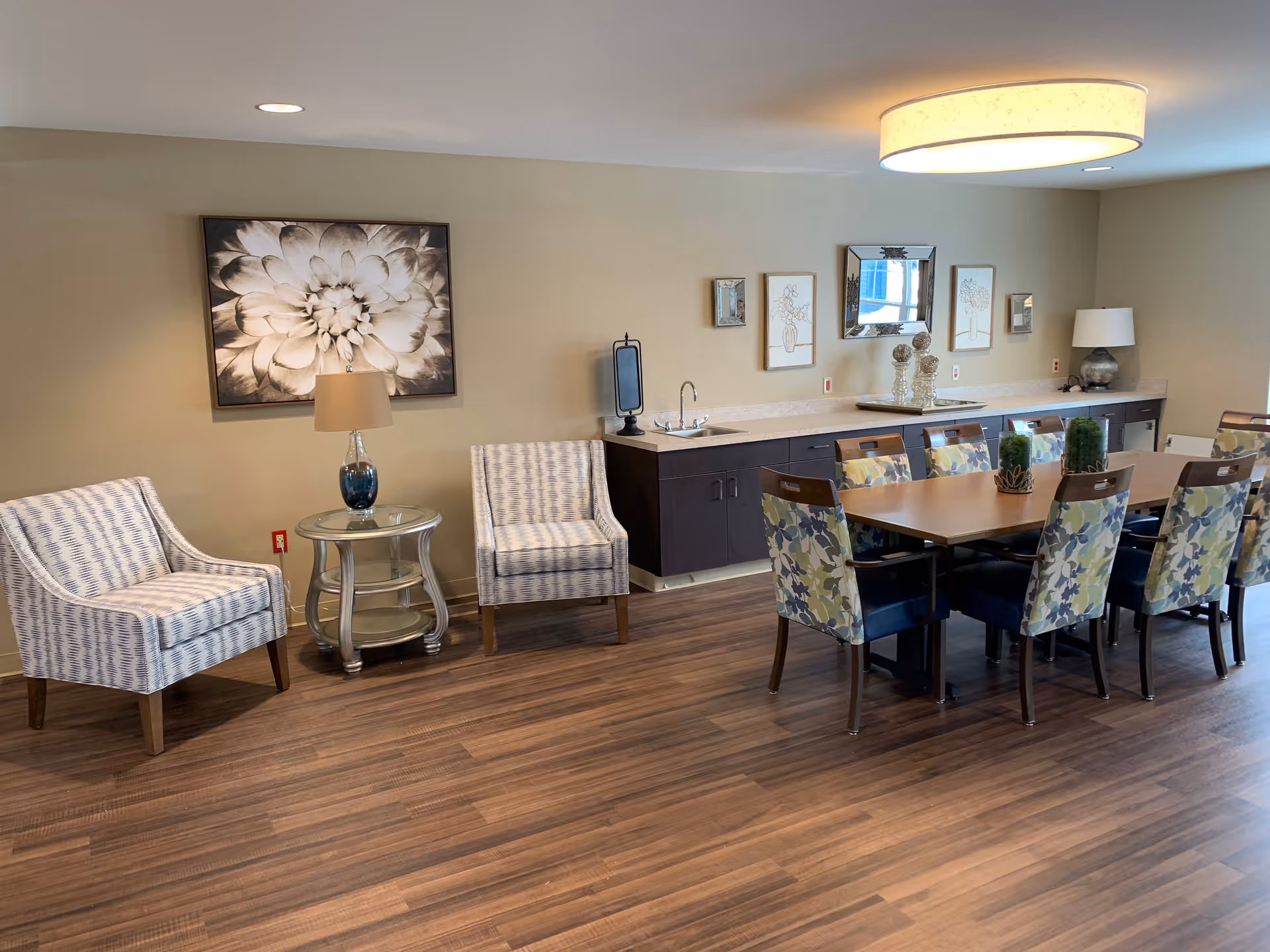 Well-lit communal dining and lounge room with a long wooden table and patterned chairs, two armchairs by a side table, and a countertop with a sink and decor.
