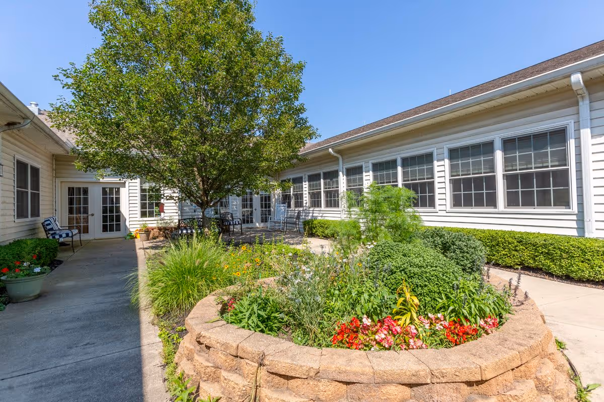 Outdoor courtyard area at Grand Victorian of Zionsville featuring a circular raised flower bed with various plants and flowers, surrounded by a concrete walkway. There are chairs and tables under a tree, and the building with multiple windows and white siding encloses the courtyard.