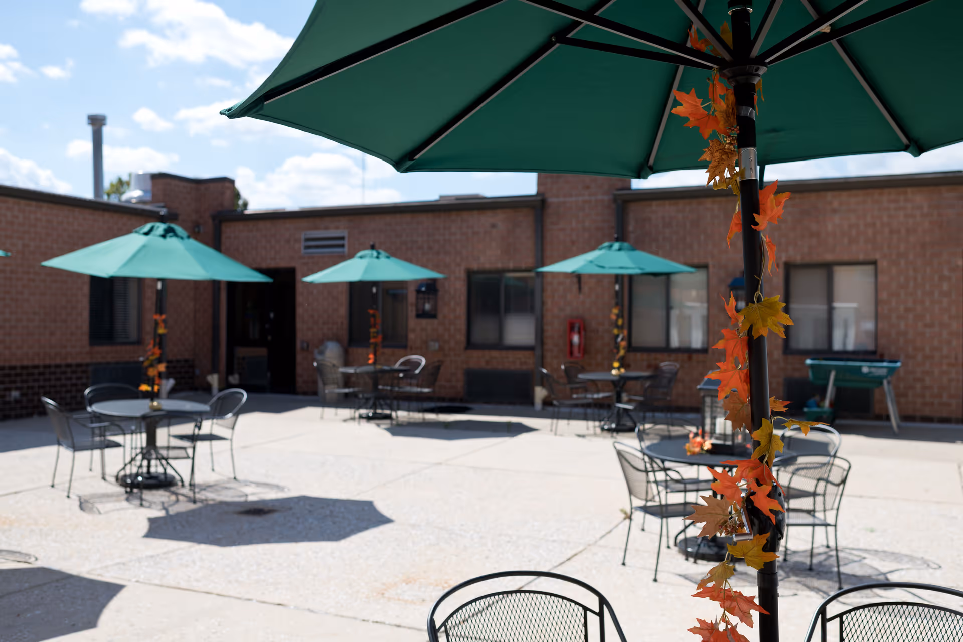 Outdoor courtyard area with several round metal tables and chairs, each shaded by large green umbrellas. The umbrellas are decorated with autumn-colored leaves. The courtyard is surrounded by a brick building under a partly cloudy sky.