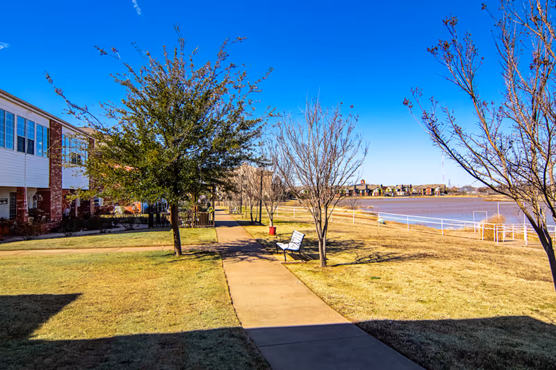 Outdoor walkway lined with trees and a bench on the right side, adjacent to a body of water. On the left side, there are residential buildings with windows and brick accents under a clear blue sky.