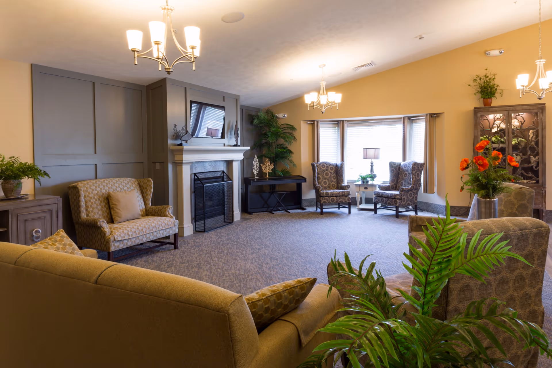 A cozy living room with beige walls and carpeted floor featuring a fireplace with a TV mounted above it. The room is furnished with multiple patterned armchairs, a sofa, a small table with a lamp near a large window, and several green plants. There are three ceiling light fixtures providing warm lighting.