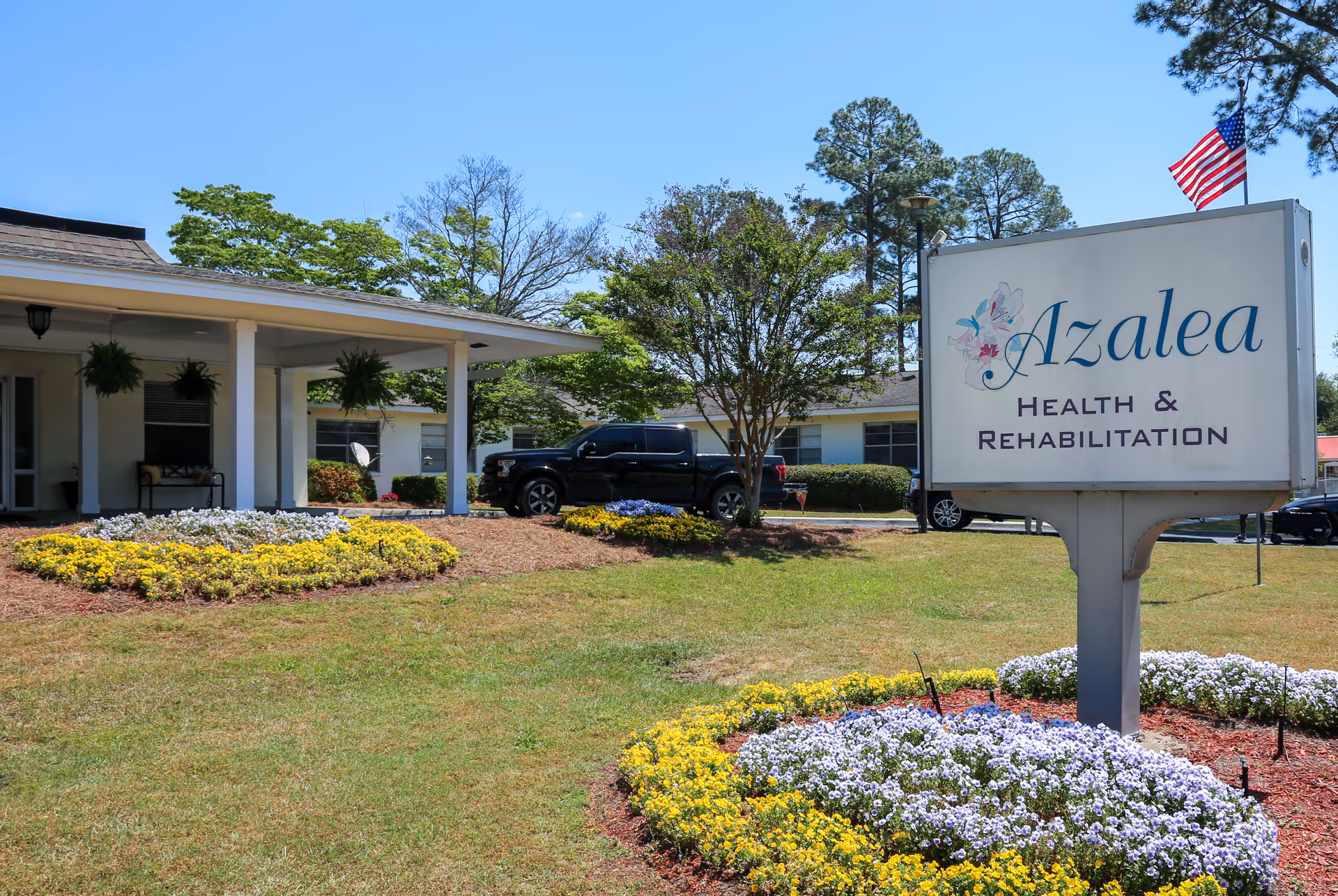 Exterior view of Azalea Health & Rehabilitation facility with a large sign displaying the facility name. The building is single-story with a covered entrance and hanging plants. There are flower beds with yellow and white flowers in the foreground, a black pickup truck parked near the building, and an American flag flying on a pole behind the sign.