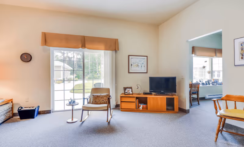 A bright living room with a sliding glass door leading outside. The room has a beige carpet and walls, a wooden TV stand with a flat-screen TV, a framed picture on the wall, a chair with a cushion, and a small round side table. There is an adjacent room visible through an open doorway with a window and a wooden chair.