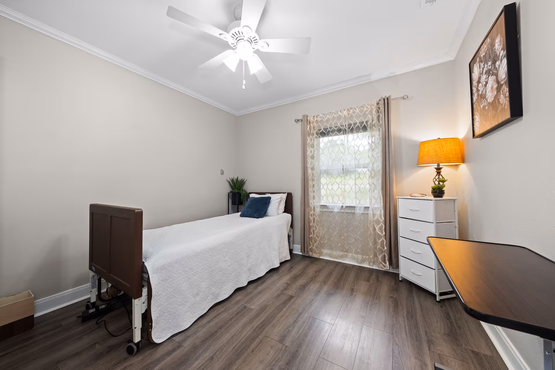 A simple, clean bedroom in a senior care facility with a single bed covered in a white quilt, a blue pillow, a small white dresser with a lamp and plant on top, a window with sheer patterned curtains, a ceiling fan with lights, and a wooden overbed table.