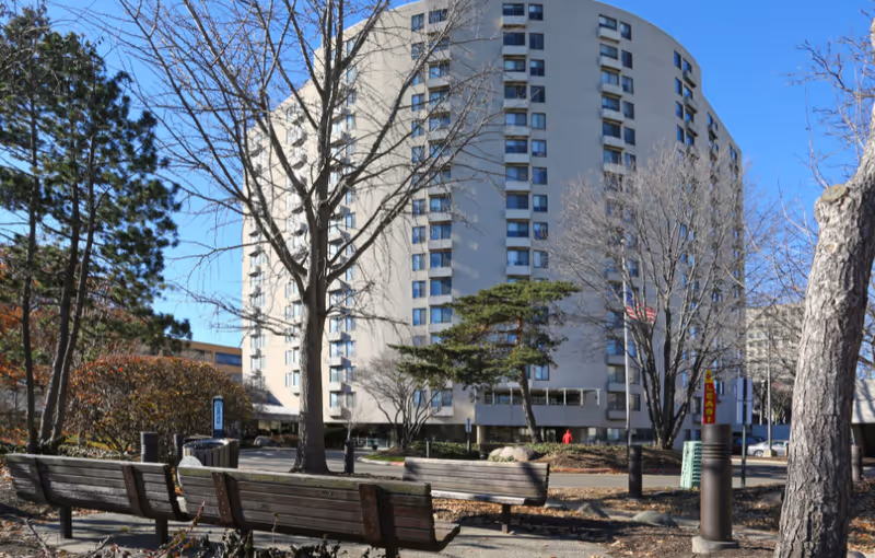 Outdoor view of a tall, multi-story residential building with several windows, surrounded by leafless trees and benches in the foreground under a clear blue sky.