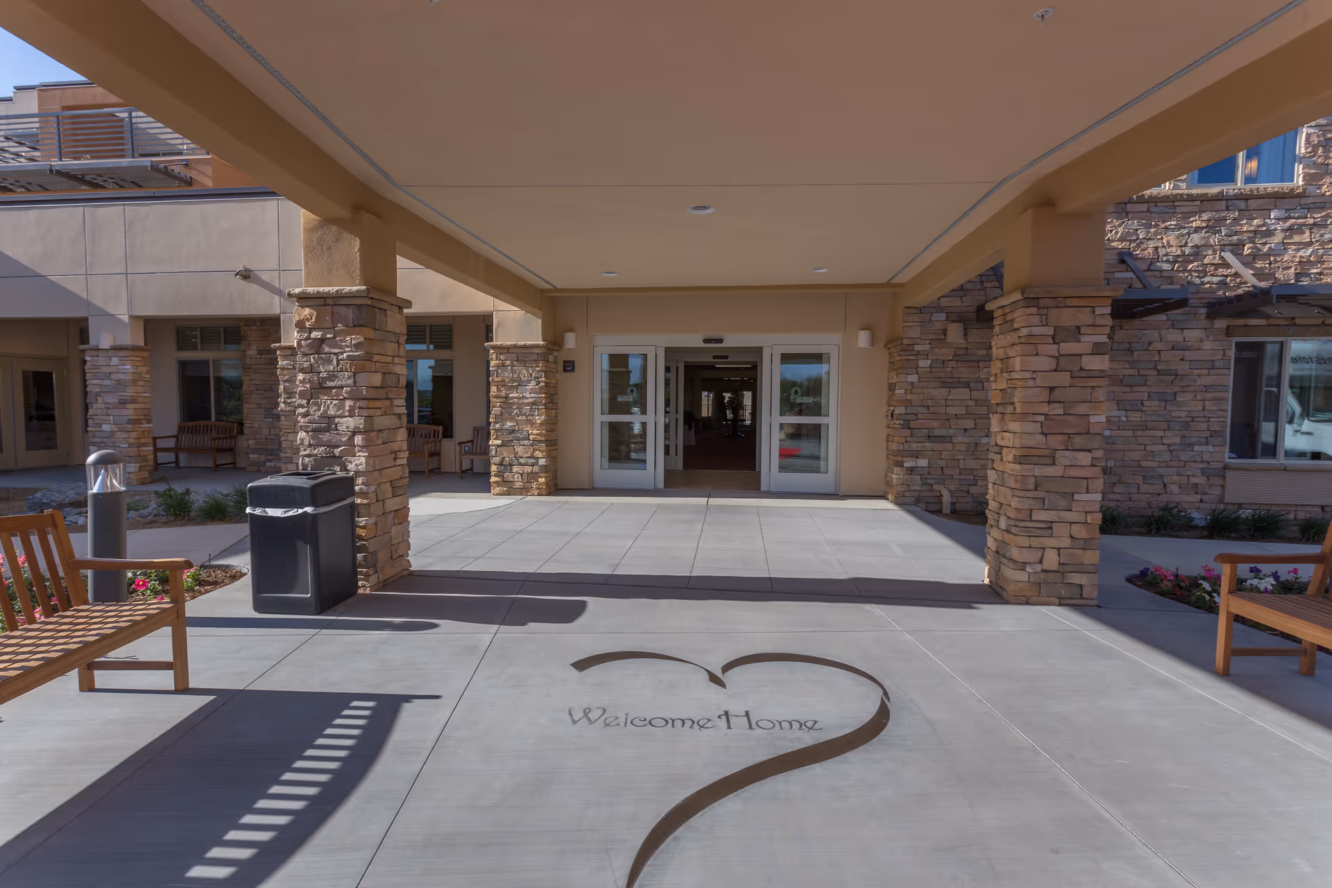 Front entrance of a senior living community with stone pillars, a covered walkway, benches, and a decorative 'Welcome Home' design on the pavement.