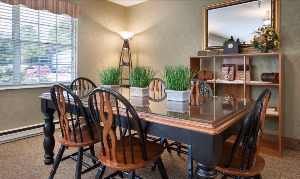 A sunlit dining room with a glass-topped wooden table surrounded by wooden chairs, potted grass centerpieces, a bookshelf and a window.