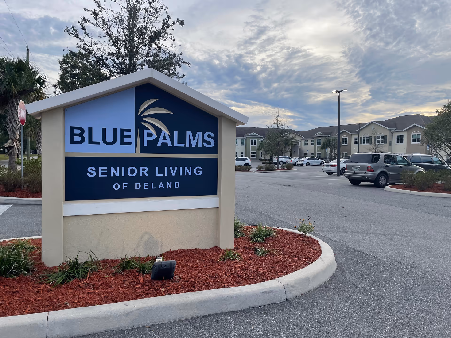 Entrance sign reading "Blue Palms Senior Living of Deland" with the facility buildings and parking lot behind it under a cloudy sky.