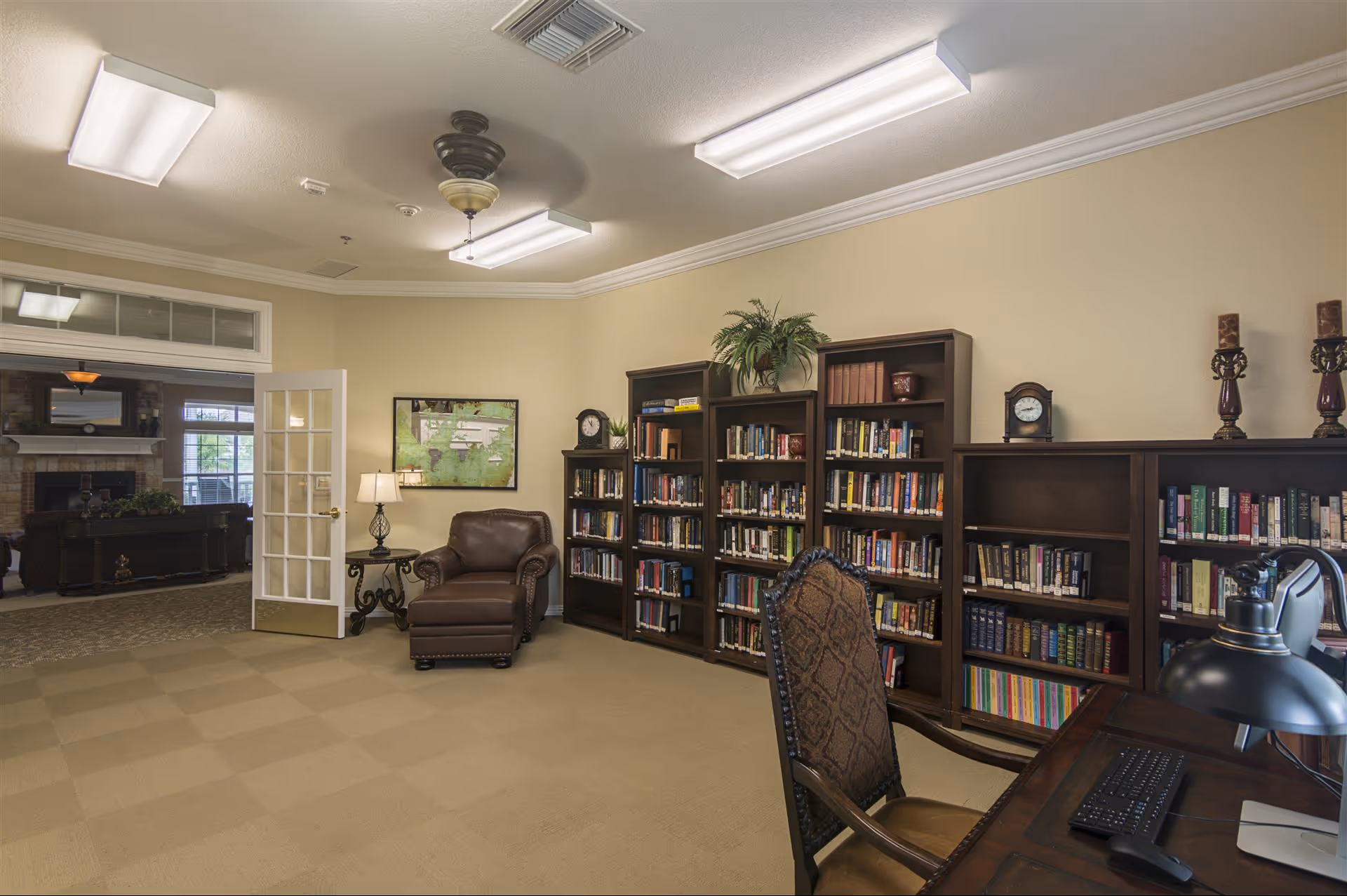 A cozy library or reading room in an assisted living facility with beige walls and carpet. There are several dark wood bookshelves filled with books, a comfortable brown leather armchair with an ottoman, a side table with a lamp, and a desk with a computer keyboard and lamp. A glass-paneled door opens to another room with a fireplace and large windows.