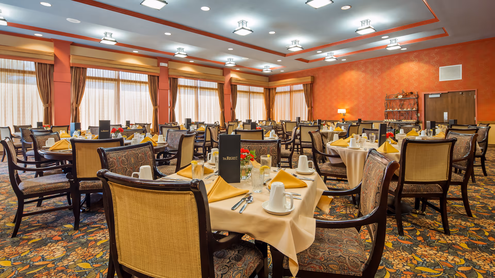 A spacious dining room with multiple round tables covered with beige tablecloths, each set with yellow folded napkins, white cups, glasses of water with lemon, and silverware. The chairs have dark wooden frames with patterned upholstery. Large windows with sheer curtains and orange drapes line one side of the room, and the walls are painted orange with a subtle pattern. The ceiling has recessed lighting fixtures.