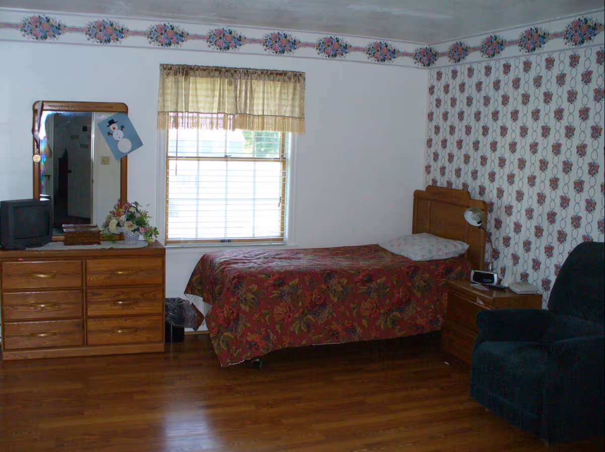A bedroom with a single bed covered with a floral bedspread, a wooden dresser with a mirror and a small TV on top, a window with a beige curtain, a nightstand with a lamp and clock, and a dark upholstered armchair. The walls have floral wallpaper and a floral border near the ceiling.