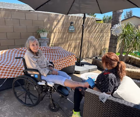 An elderly woman in a wheelchair is sitting outside under a large umbrella while a caregiver wearing gloves tends to her foot. They are in a patio area with a brick wall, plants, and outdoor furniture.