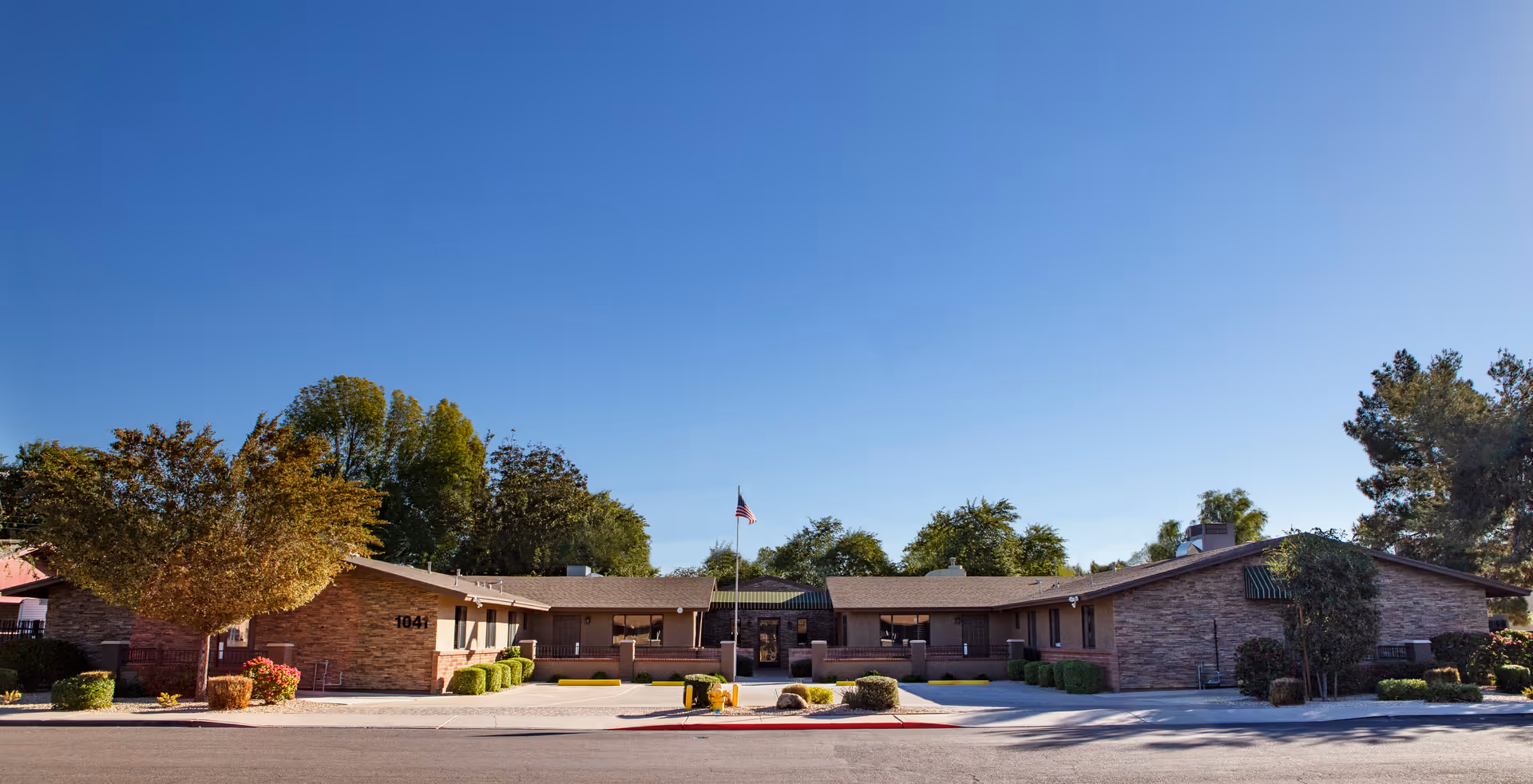 Front exterior view of a single-story brick building with a central entrance, surrounded by trees and shrubs under a clear blue sky. An American flag is displayed on a flagpole in front of the entrance.