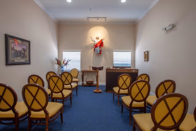 Small chapel room with rows of wooden chairs with yellow cushions facing a podium and a statue of Jesus on the wall between two windows. There is a framed picture on the left wall and a small table with flowers beneath the windows.