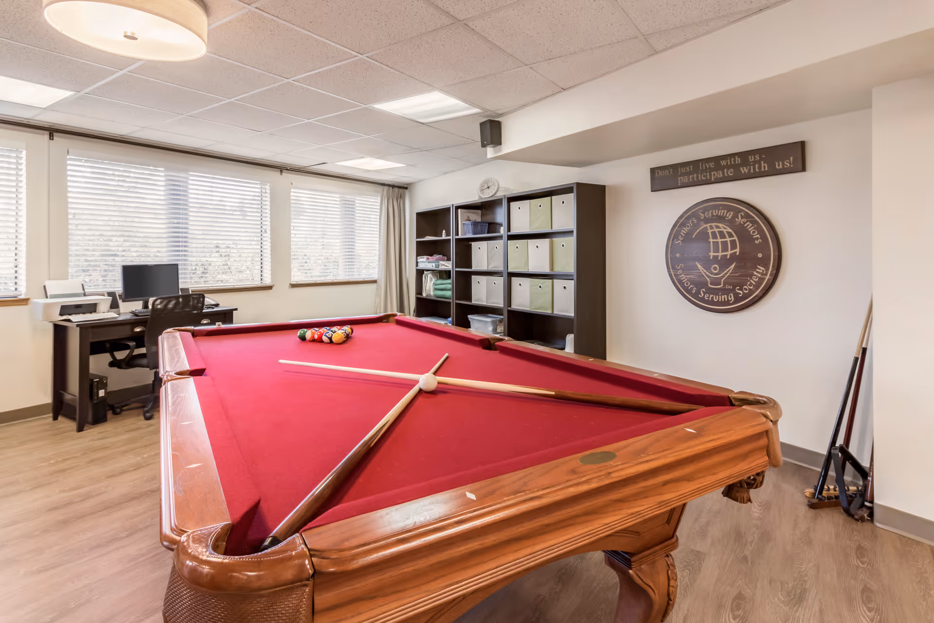 A bright interior recreation room featuring a red-felt pool table with cues, shelving, and a desk with a computer by the windows.