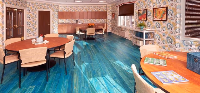 Communal dining room with round wooden tables and beige chairs on vibrant turquoise wood floors, patterned wallpaper, and windows with blinds.