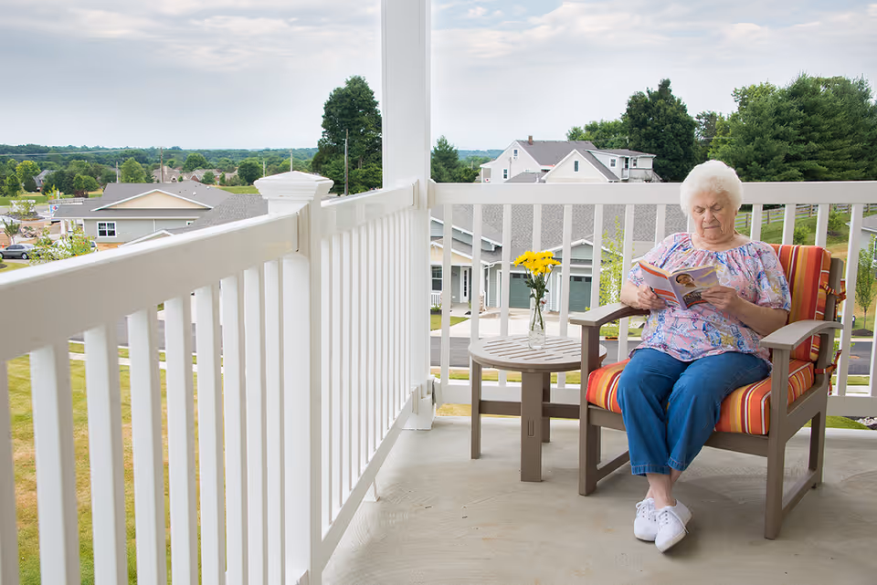 An elderly woman sitting on a striped cushioned chair on a balcony, reading a book. Next to her is a small round table with a vase of yellow flowers. The balcony has white railings and overlooks a neighborhood with houses and trees under a cloudy sky.