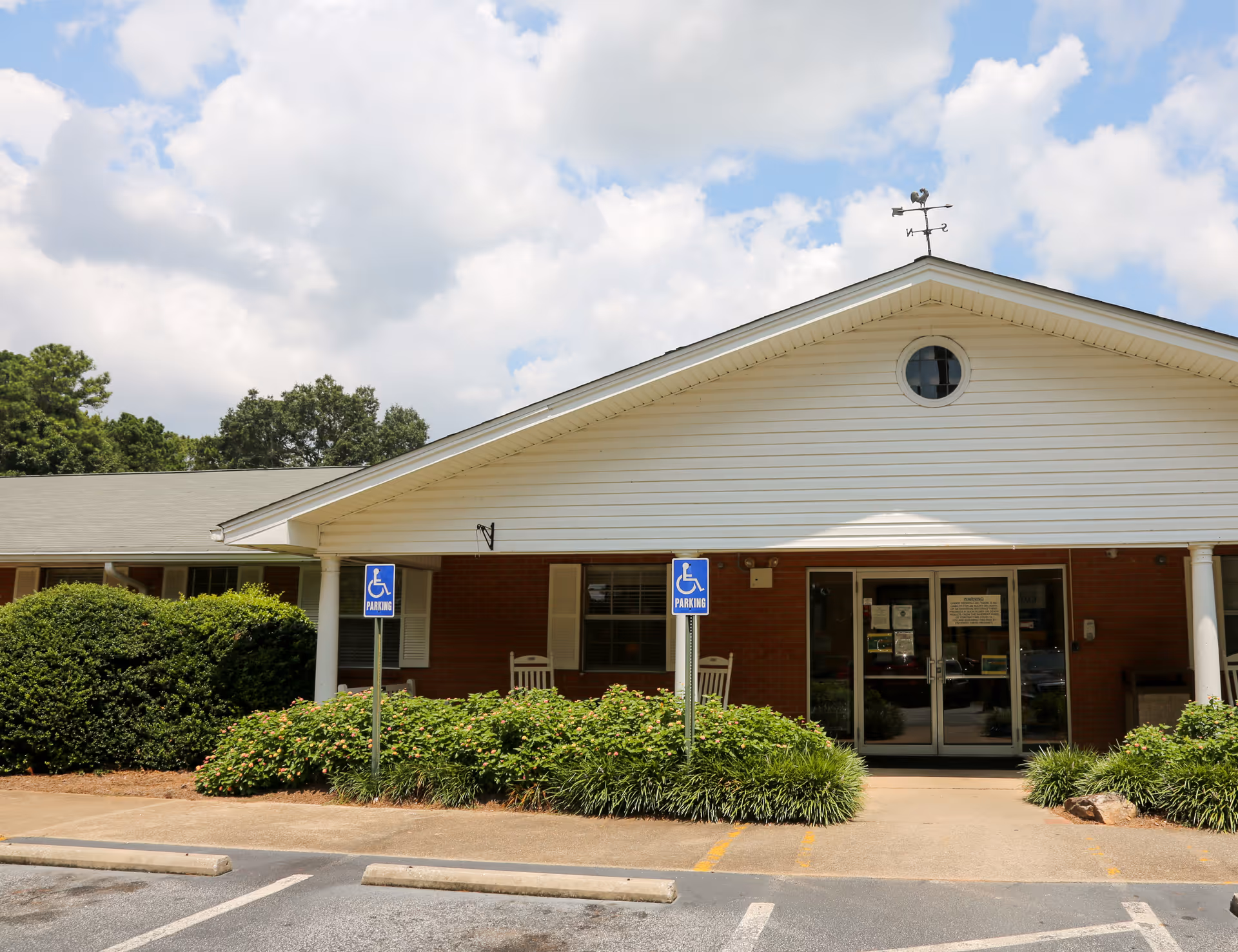 Front entrance of a single-story brick and white-sided senior living facility with a covered porch, handicap parking signs, and landscaping.