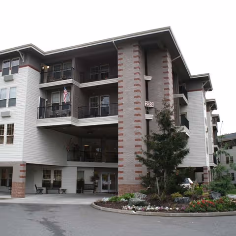 Exterior view of a multi-story senior living facility building with balconies, an American flag, and a landscaped area with flowers and shrubs near the entrance.