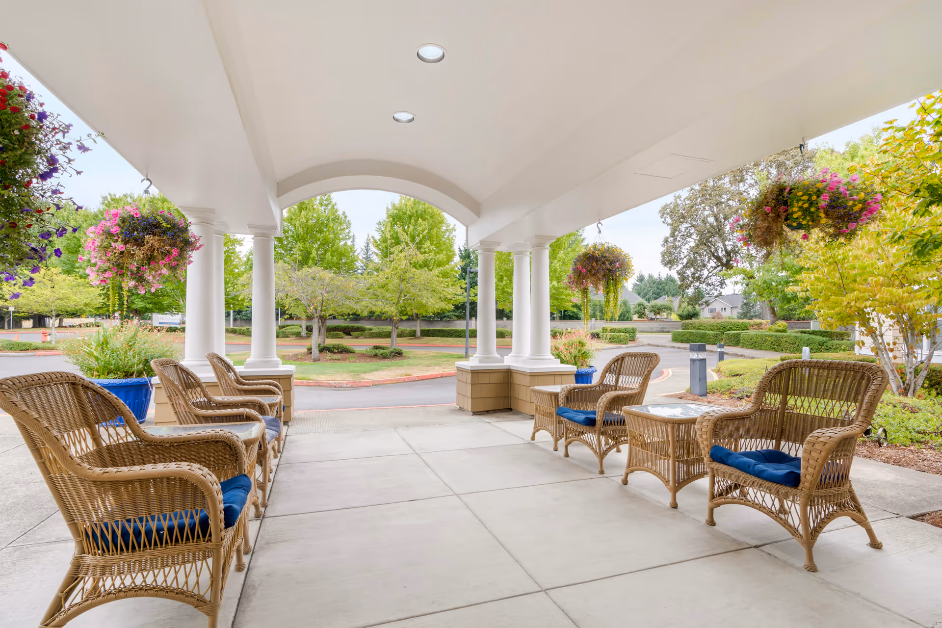 Covered outdoor seating area with wicker chairs and tables, hanging flower baskets, and a view of a landscaped garden and trees in the background.