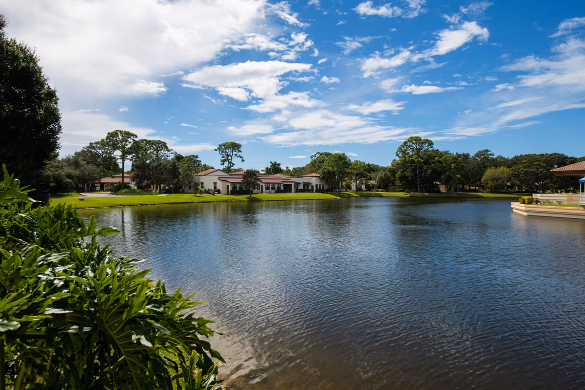 A serene outdoor view of a pond with clear blue water surrounded by green grass, trees, and shrubs. In the background, there are single-story buildings with red-tiled roofs under a partly cloudy blue sky.