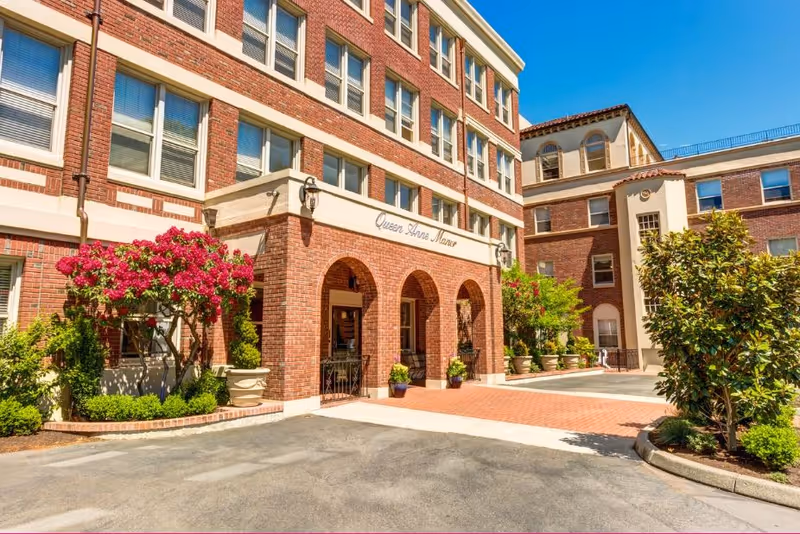 Brick exterior and arched main entrance of Queen Anne Manor with potted plants and flowering shrubs under a clear blue sky.