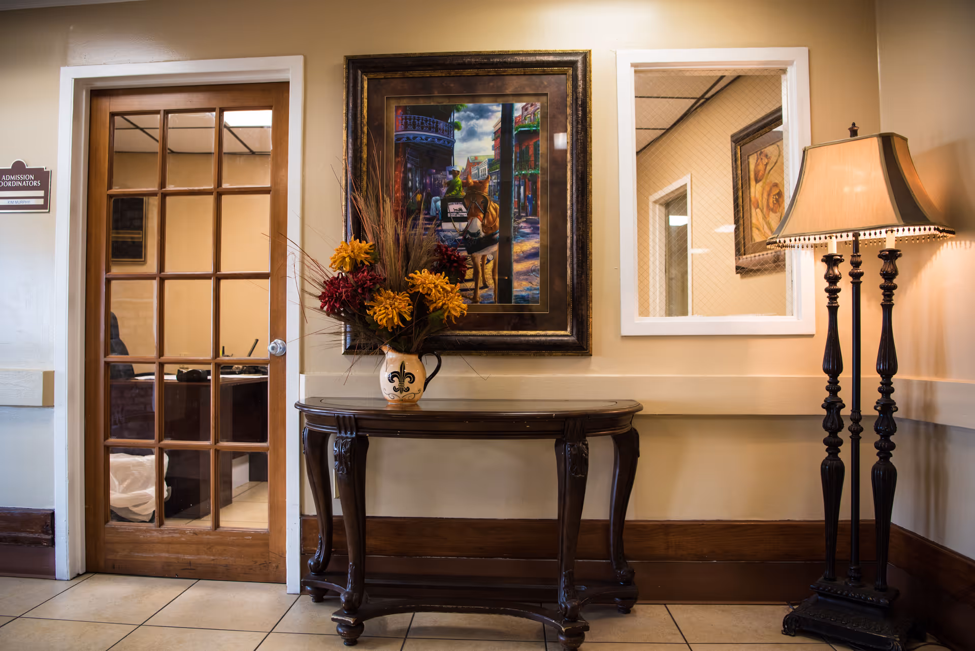 A decorated hallway interior with a wooden console table holding a floral arrangement beneath a framed painting, next to a glass-paned door and a tall floor lamp.