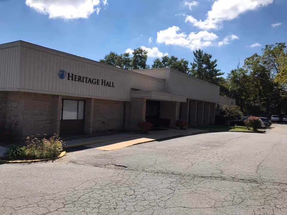 Exterior view of Heritage Hall Leesburg building with a parking area in front, surrounded by trees under a partly cloudy sky.