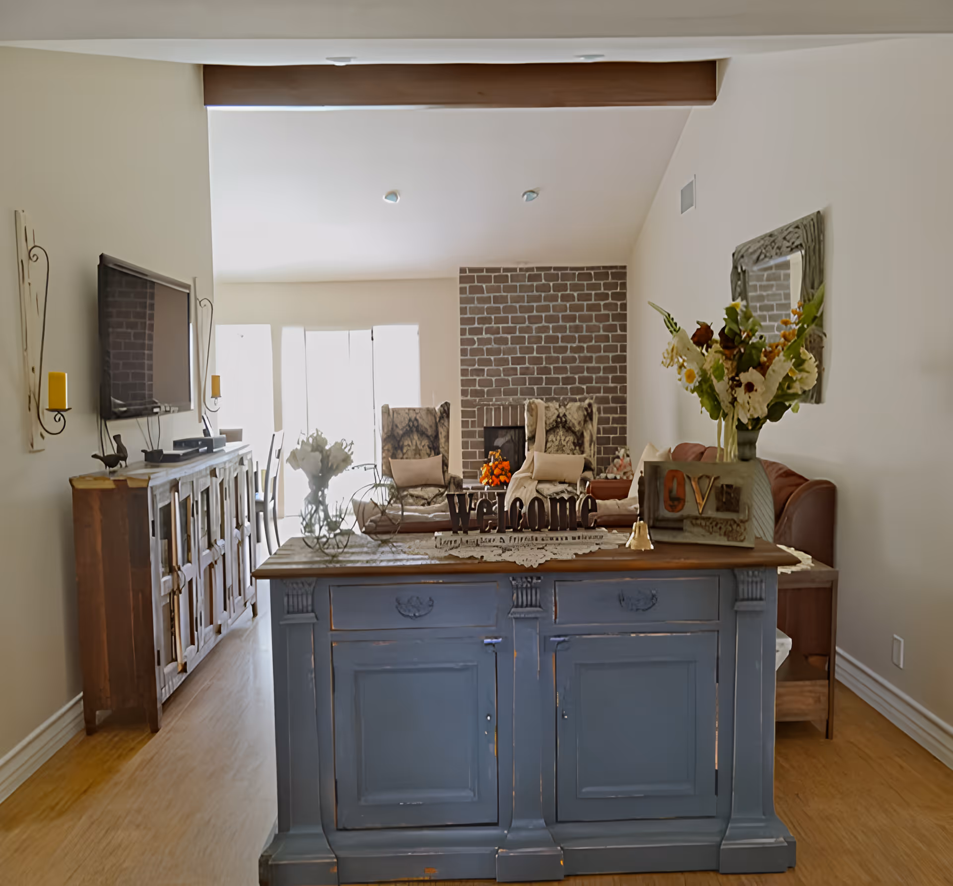 A cozy living room with a blue wooden cabinet in the foreground decorated with a vase of flowers and a 'Welcome' sign. In the background, there are two patterned armchairs with cushions in front of a brick fireplace. To the left, a wooden sideboard with a mounted flat-screen TV and yellow candles on the wall. The room has light-colored walls and wooden flooring.