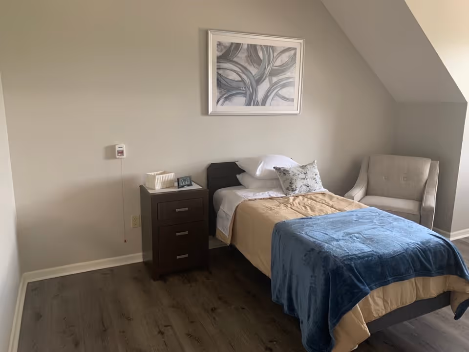A simple bedroom with a single bed, nightstand, armchair, and abstract wall art.