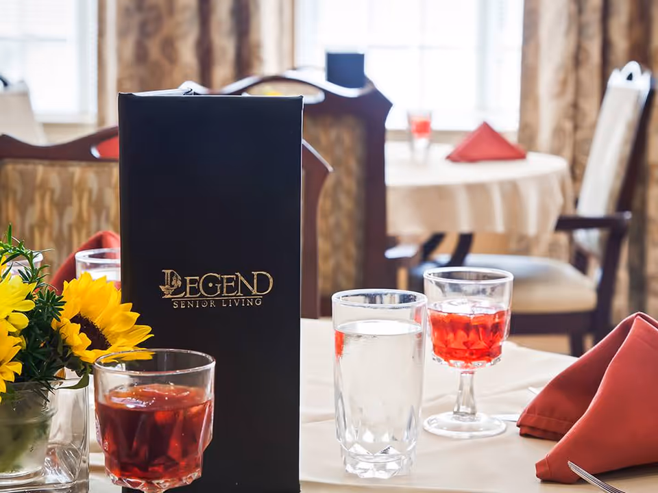 A dining table set with a black menu labeled 'Legend Senior Living', two glasses with red beverage and one glass of water, a small vase with yellow sunflowers, and red folded napkins. In the background, there are other tables and chairs with similar settings in a well-lit dining room.