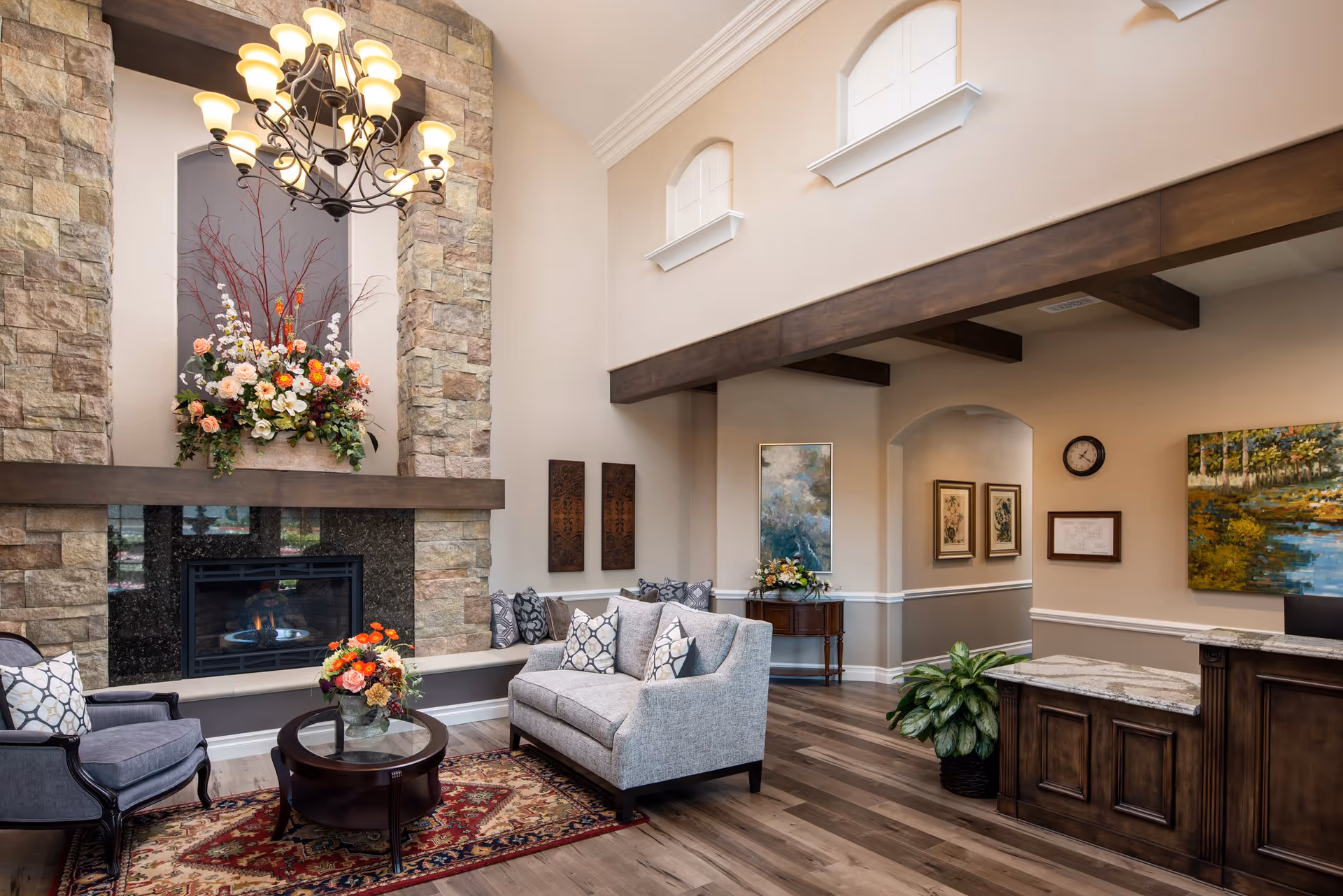 A cozy living room area in a senior living facility featuring a stone fireplace with a floral arrangement on the mantel, a chandelier overhead, a gray loveseat with patterned pillows, a gray armchair, a round glass coffee table with a flower vase, wooden flooring, and decorative paintings and plants around the room.