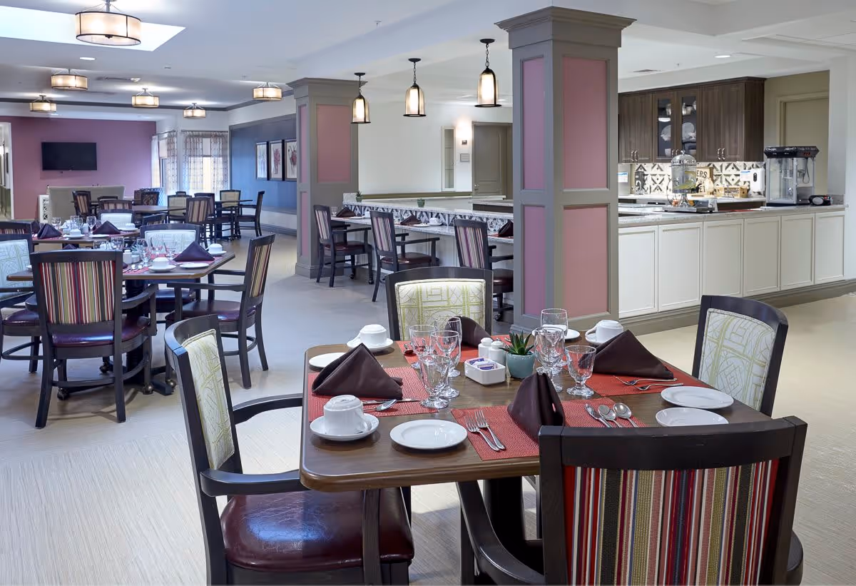 A spacious dining room with multiple tables set for a meal, featuring plates, glasses, cups, and folded napkins. The room has a modern design with striped upholstered chairs, pendant lights hanging from the ceiling, and a kitchen area with cabinets and a popcorn machine in the background.