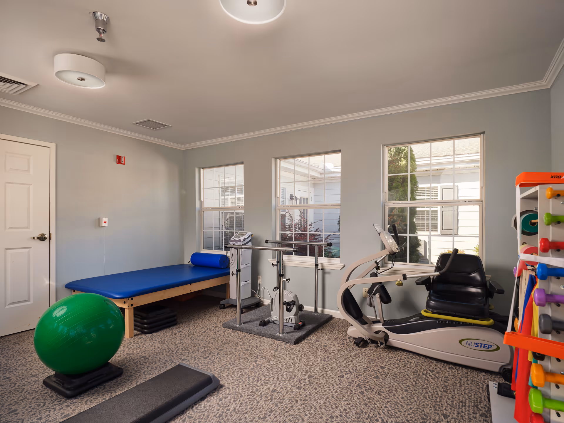 A small fitness or physical therapy room with exercise equipment including a green exercise ball, a blue padded therapy table, parallel bars for walking exercises, a recumbent exercise bike, and a rack holding various colorful dumbbells. The room has light gray walls, two large windows letting in natural light, and a patterned carpeted floor.