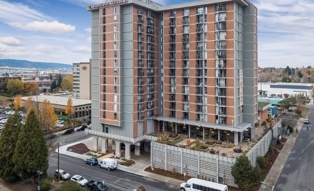A multi-story senior living facility named Calaroga Terrace with a brick and gray exterior, surrounded by parking areas, trees, and nearby buildings under a partly cloudy sky.