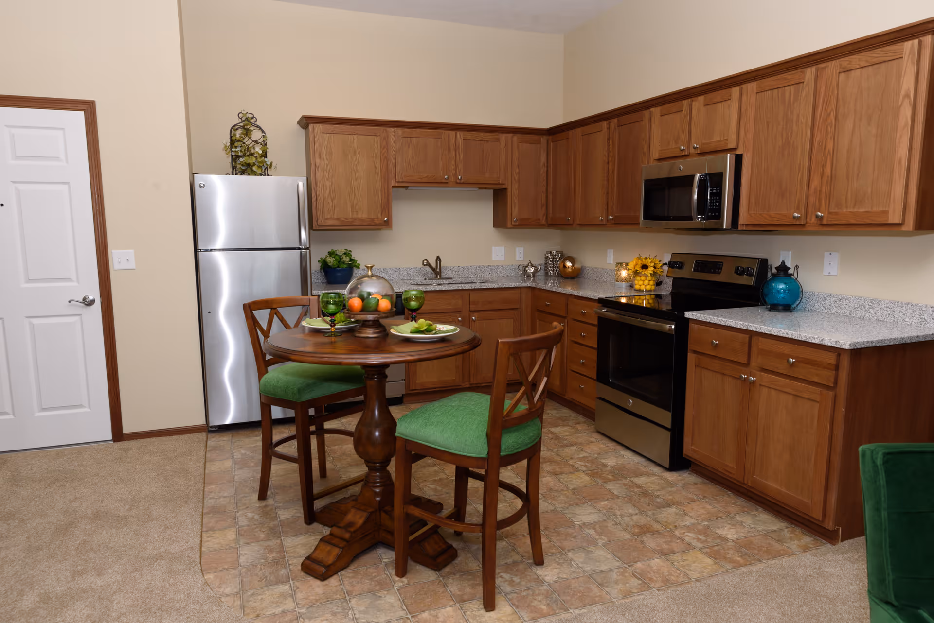 A kitchen area with wooden cabinets, a stainless steel refrigerator, stove, and microwave. There is a small round wooden table with two chairs that have green cushions. The floor is tiled in the kitchen area and carpeted in the adjacent space.