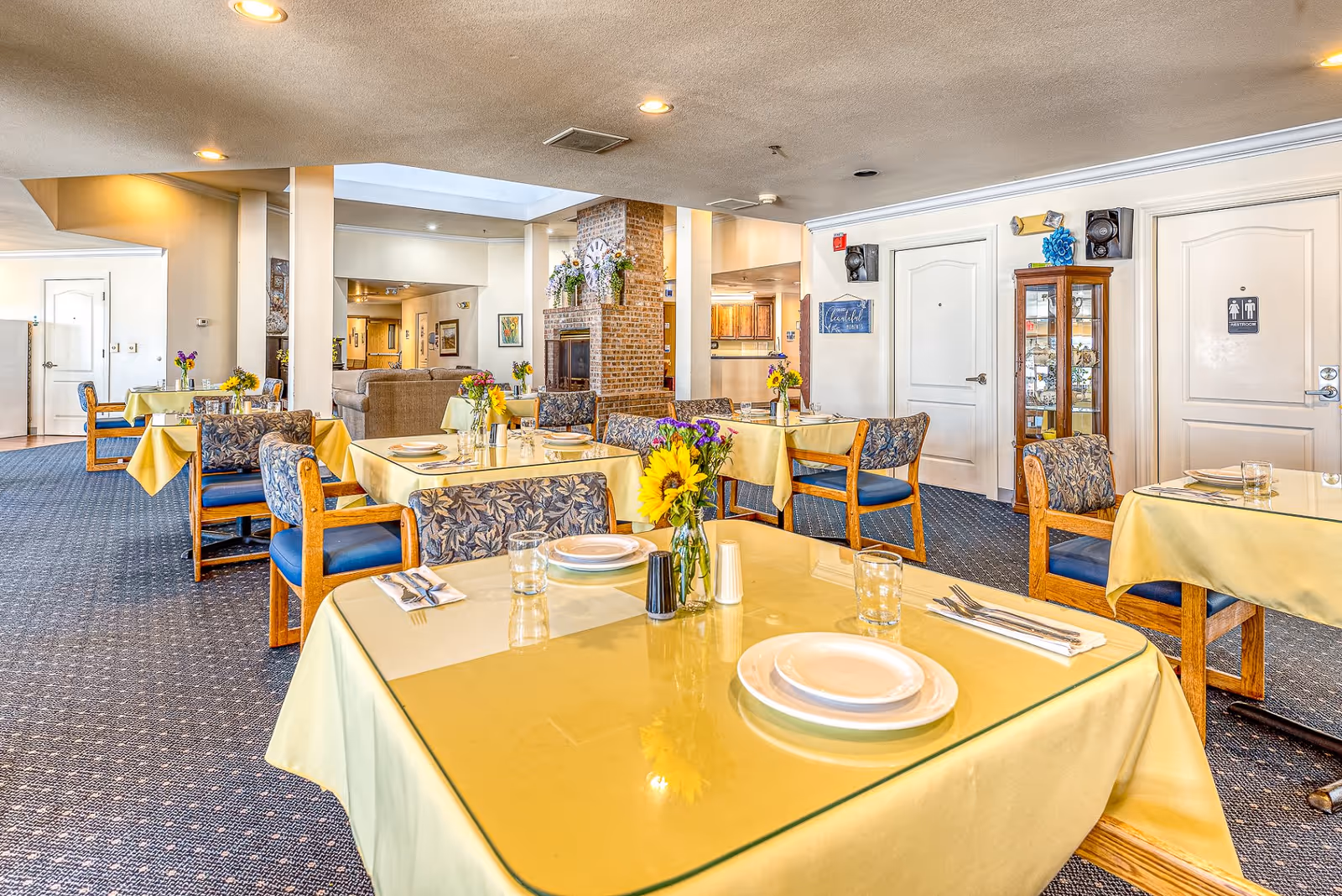 A bright dining room with several tables covered in yellow tablecloths and set with plates, glasses, and silverware. Each table has a small vase with colorful flowers. The room has carpeted floors, patterned chairs, and a brick fireplace in the background. There are doors on the right side, one with a restroom sign. The overall atmosphere is clean and inviting.