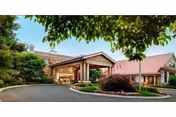 Exterior view of a senior living facility named Lima Estates featuring a covered entrance with a circular driveway, surrounded by landscaped greenery and trees under a clear sky.