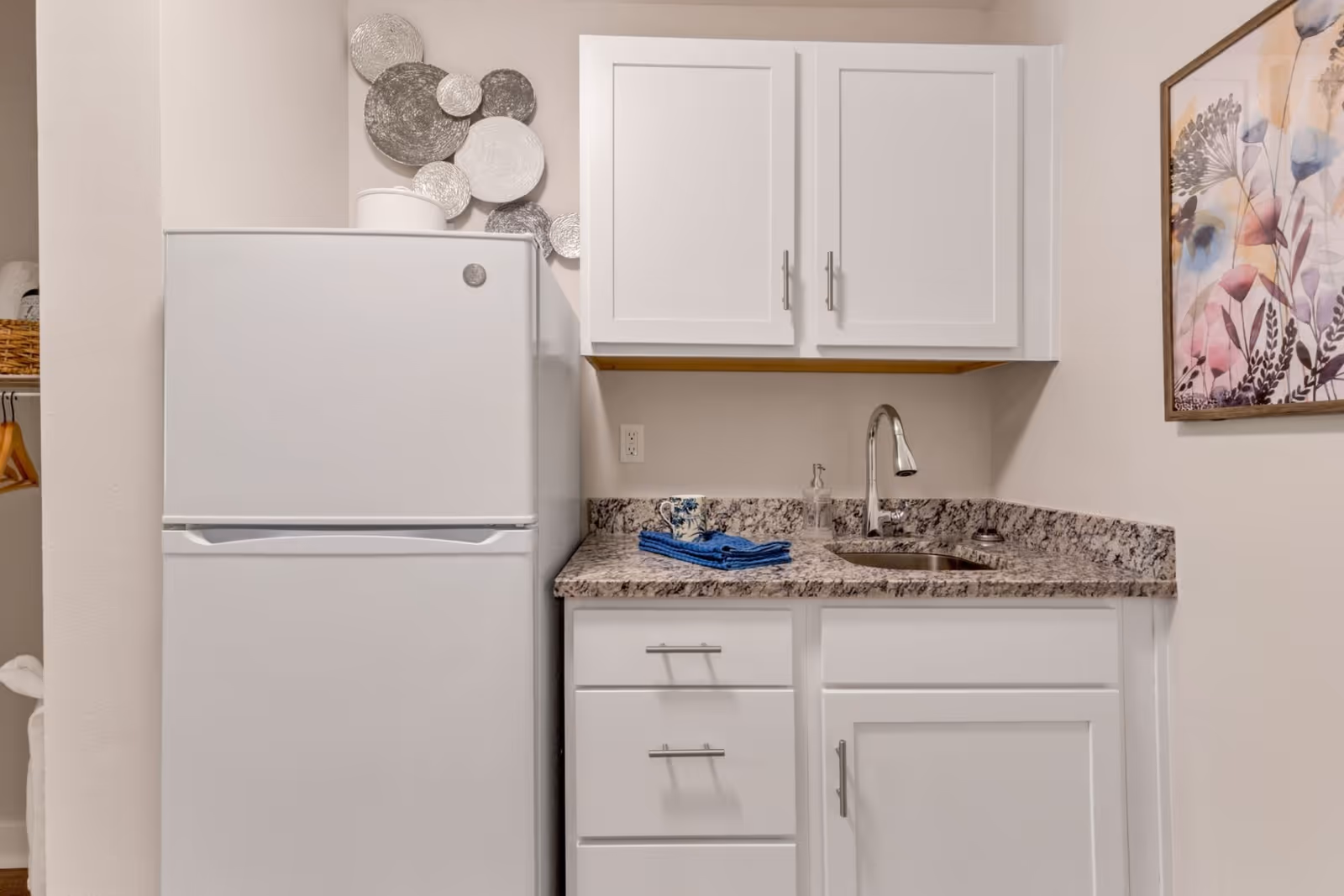 A small kitchenette area featuring a white refrigerator, white cabinets with silver handles, a granite countertop with a built-in sink and faucet, and a few blue towels placed on the counter. There is a decorative wall art piece with circular metallic plates above the refrigerator and a framed floral painting on the adjacent wall.