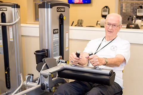 An elderly man using a seated leg curl exercise machine in a fitness room. He is wearing glasses and a white polo shirt, and appears to be smiling while exercising. The background shows other exercise equipment and a television mounted on the wall.