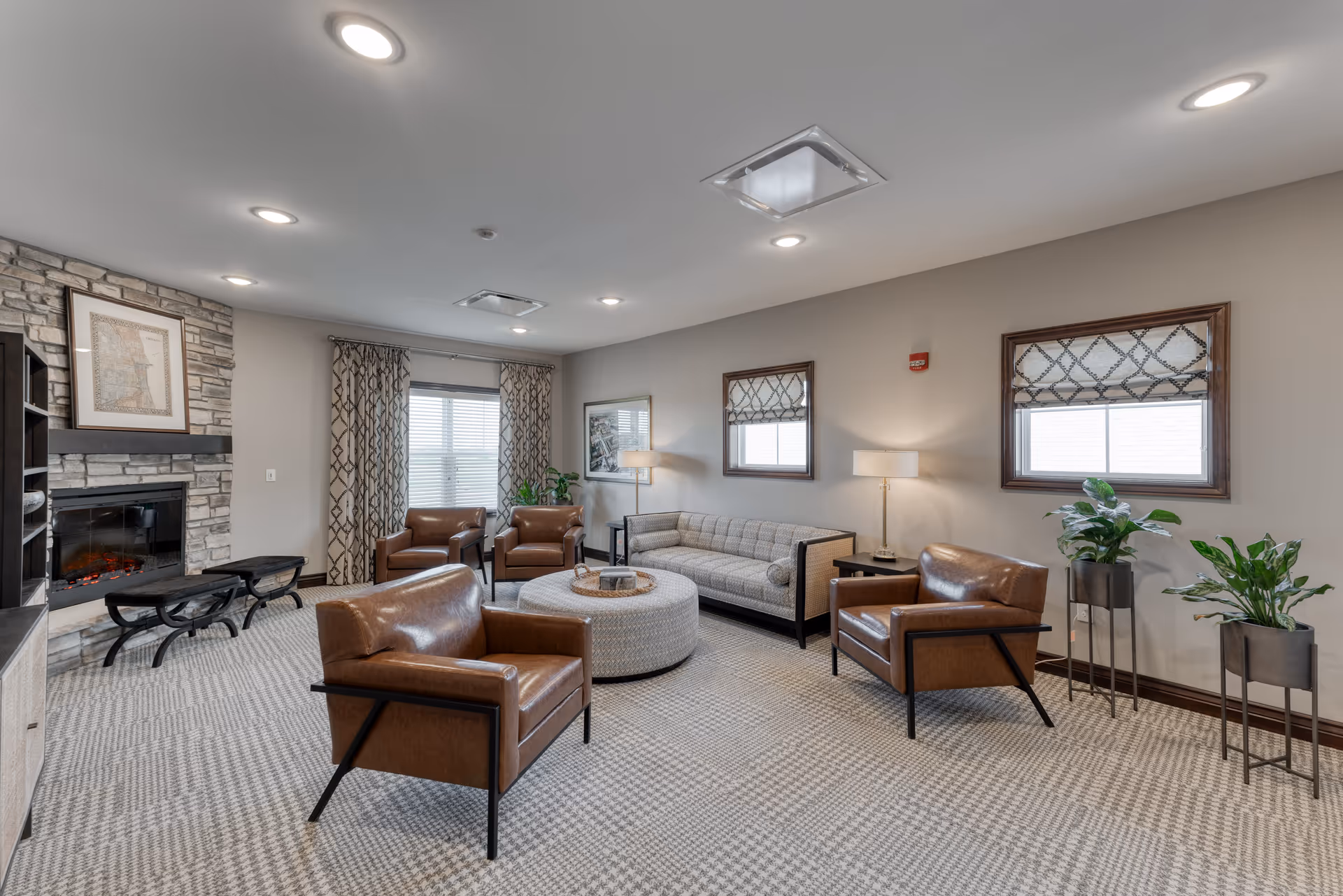 A bright senior living common room with leather armchairs, a sofa, a round ottoman, a fireplace, and potted plants.