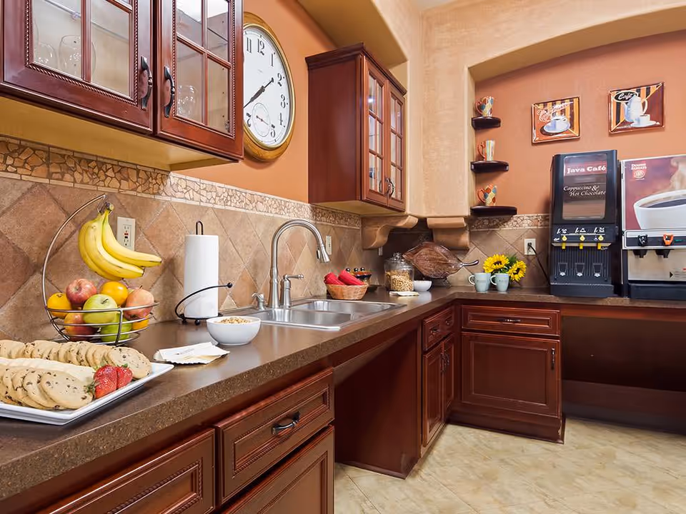 A kitchen area with dark wood cabinets, a double sink, and a countertop displaying a basket of fruit, a plate of cookies with strawberries, a bowl of cereal, and a paper towel holder. On the right side, there is a coffee and hot chocolate dispenser with two mugs and a small vase of sunflowers. The walls are painted warm tones with decorative tiles and a large clock above the sink.