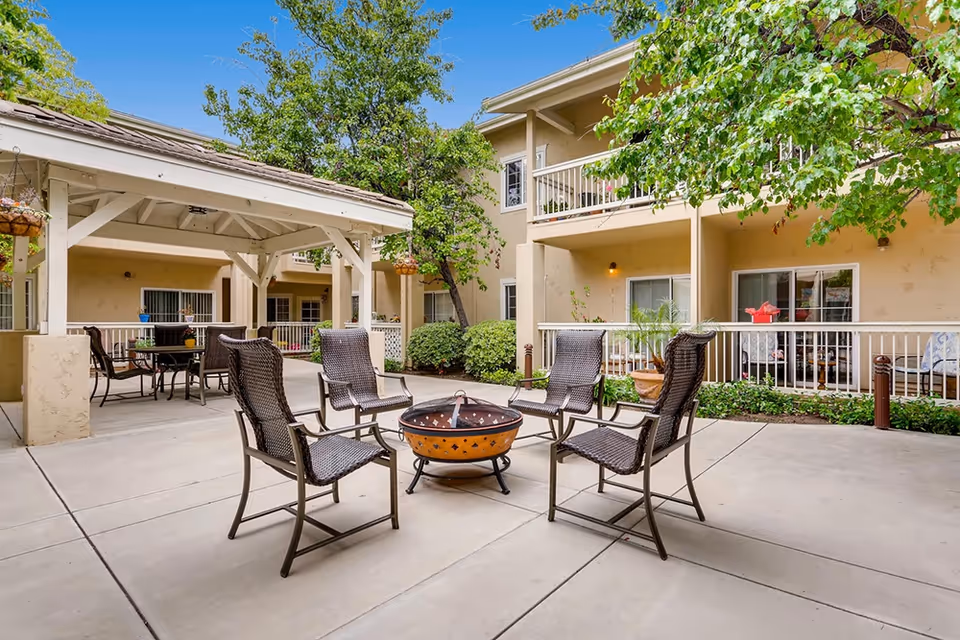 Sunny courtyard at a senior living facility with a fire pit surrounded by wicker chairs, a covered patio area, and two-story balconies.