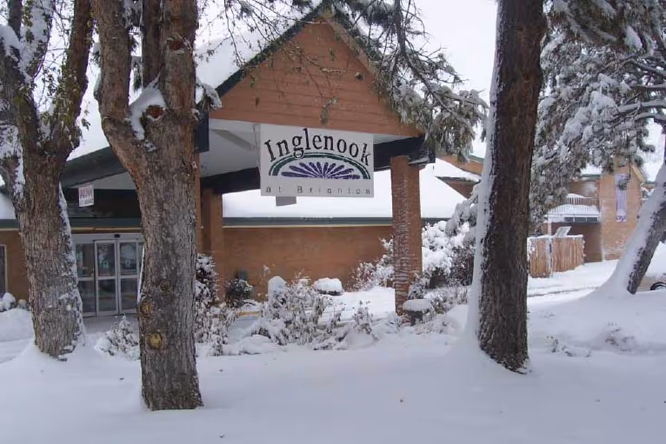 Snow-covered entrance of Inglenook At Brighton facility with trees and bushes blanketed in snow in the foreground.