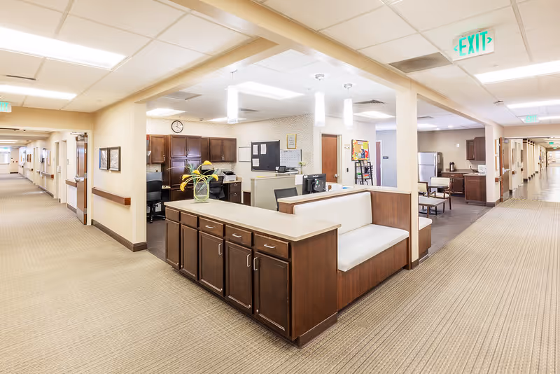 Interior view of a senior living facility hallway with a reception desk area in the center. The desk has dark wood cabinets and a white cushioned bench. There are two long corridors extending to the left and right with doors along the walls. The area is well-lit with ceiling lights and has beige walls and carpeted floors. An exit sign is visible on the ceiling.