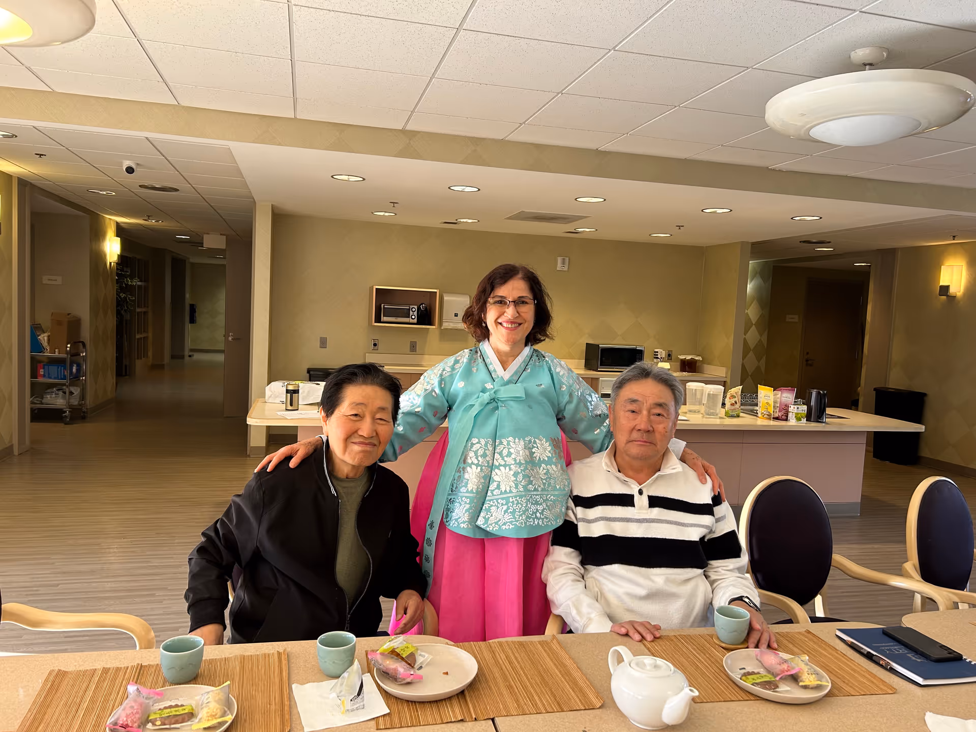 Three people in a senior living facility dining area. Two elderly individuals are seated at a table with tea cups and plates of snacks, while a woman in traditional Korean attire stands behind them with her arms around their shoulders, smiling. The room has a kitchen counter with a microwave and various items in the background.