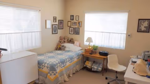 A small, cozy bedroom with a single bed covered by a blue and yellow patterned quilt. Several teddy bears are placed on the bed near the pillows. The walls are decorated with multiple framed pictures. There are two windows with white blinds letting in natural light. A wooden side table next to the bed holds a lamp, a plant, and a radio. A white chair with wheels is positioned near a desk with various items on it.